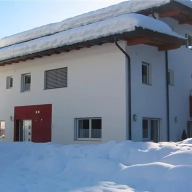 A modern, white house with a red entrance area, surrounded by snow-covered terrain. The roof edge is covered with snow, and a clear sky can be seen.