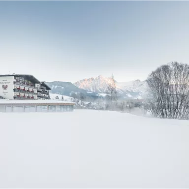 A snow-covered landscape with a hotel in the foreground and mountains in the background. The sky is clear and the scene radiates calmness and cold.