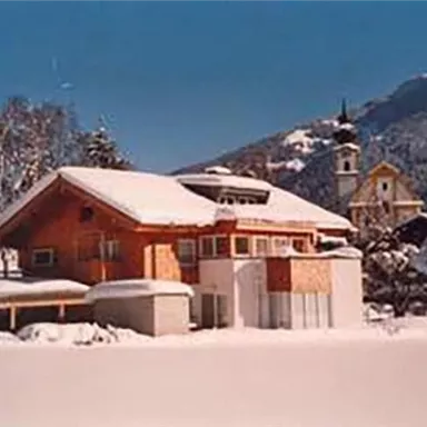 Ein gemütliches Holzhaus im Schnee, umgeben von einer winterlichen Landschaft. Im Hintergrund sind Berge und eine Kirche zu sehen.