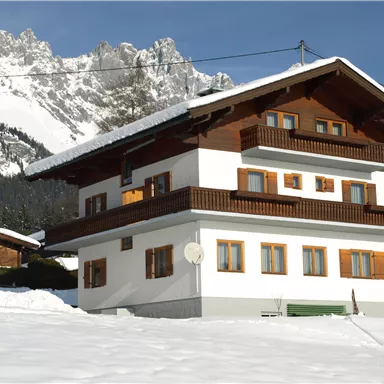 Ein schönes, zweigeschossiges Chalet in den Alpen, umgeben von Schnee. Im Hintergrund erheben sich majestätische Berge unter einem klaren Himmel.