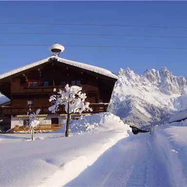 Ein traditionelles Holzhaus im Schnee, umgeben von majestätischen Bergen. Der Himmel ist klar und blau.