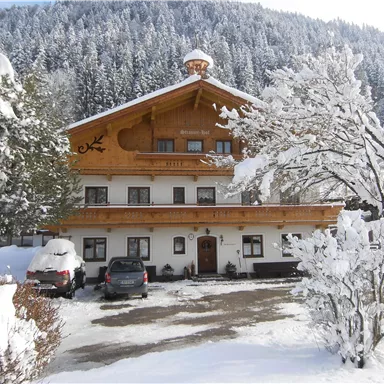 Ein alpenländliches Haus im Winter mit schneebedecktem Dach. Umgeben von verschneiten Bäumen und einem malerischen Bergblick.