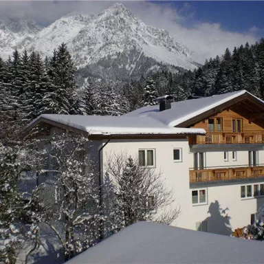 Ein schneebedecktes Haus umgeben von dichten, grünen Wäldern und majestätischen Bergen. Der Himmel ist klar und die Landschaft vermittelt eine winterliche Ruhe.