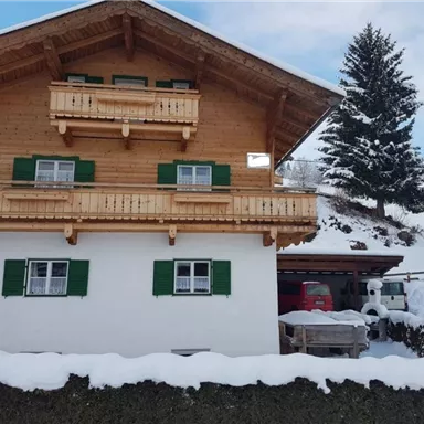 Ein gemütliches Holzhaus im Schnee mit grünen Fensterläden. Umgeben von einer winterlichen Landschaft und Tannenbäumen.