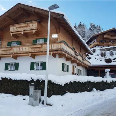 Ein charmantes Chalet im Winter mit weißem Schnee und grünen Fenstern. Im Hintergrund sind weitere Holzhäuser und schneebedeckte Bäume zu sehen.