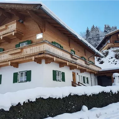 Ein traditionelles chaletartiges Haus im Schnee mit grünen Fensterläden. Die umliegende Landschaft ist winterlich und malerisch.