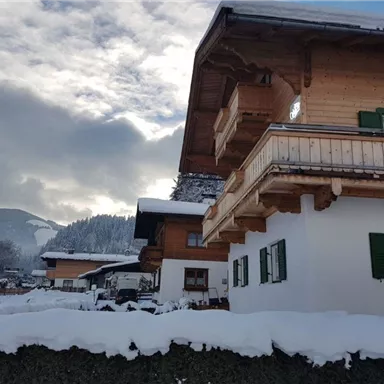 Ein malerisches Winterdorf mit schneebedeckten Dächern und einem hügeligen Hintergrund. Die Gebäude haben traditionelle Holzverzierungen und grüne Fensterläden.