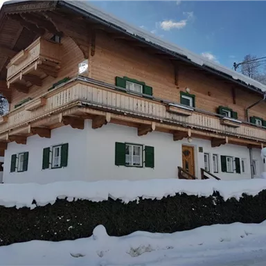 Ein charmantes Holzhaus im Winter, umgeben von Schnee. Die grünen Fensterläden und der Balkon verleihen dem Gebäude eine alpine Note.