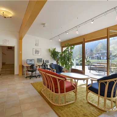 Ein helles Wohnzimmer mit großen Fenstern und Blick auf die Natur. Es gibt bequeme Möbel und einen Pflanzenakzent im Raum.