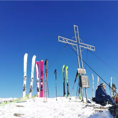 Eine Schneelandschaft mit einem Kreuz und Skiern. Zwei Personen sitzen auf dem Schnee unter einem klaren blauen Himmel.