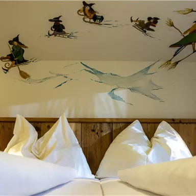 Ein gemütliches Schlafzimmer mit weißen Kissen und einer künstlerischen Wanddekoration. Die Wand zeigt Figuren, die auf Skiern unterwegs sind, und verleiht dem Raum einen märchenhaften Charme.