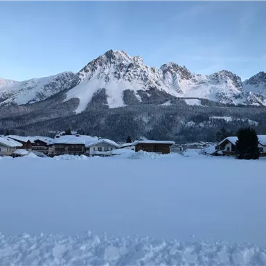 Eine verschneite Landschaft mit Bergen im Hintergrund. Die Häuser im Vordergrund sind von Schnee bedeckt.