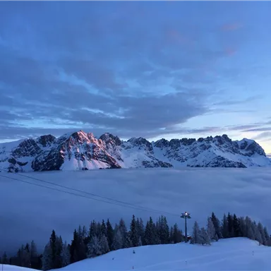 Eine majestätische Berglandschaft mit schneebedeckten Gipfeln. Darunter breitet sich eine Wolkendecke aus, während der Himmel sanft blau und bewölkt ist.