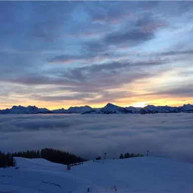 Ein atemberaubender Blick auf schneebedeckte Berge, die über einer dichten Wolkendecke liegen. Der Himmel strahlt in sanften Farben des Sonnenuntergangs.