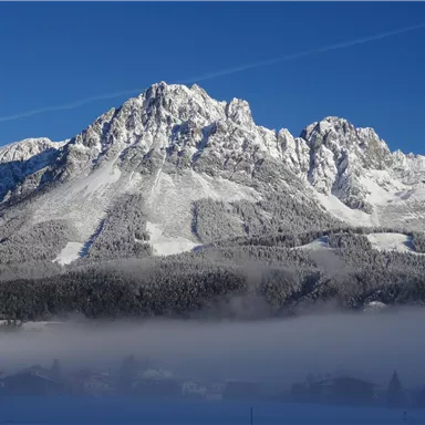 Ein majestätischer Berg mit schneebedeckten Gipfeln unter einem klaren blauen Himmel. Am Fuße des Berges liegt eine leichte Nebelschicht über der Landschaft.