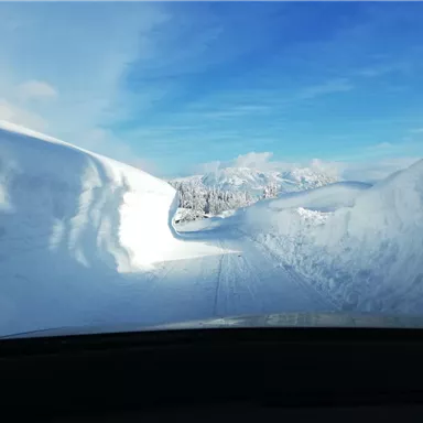 Eine schneebedeckte Straße, umgeben von hohen Schneewänden. Der Himmel ist blau und die Berge sind im Hintergrund sichtbar.
