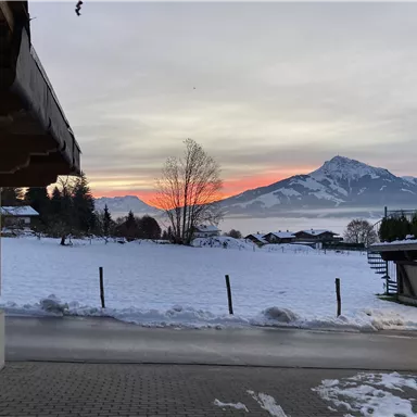 Ein winterlicher Landschaftsausblick mit schneebedeckten Feldern und Bergen im Hintergrund. Der Himmel leuchtet in sanften Farben des Sonnenuntergangs.