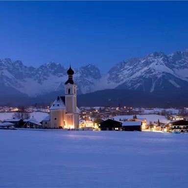 A picturesque winter landscape with snow-covered fields and a village in the background. The church glows gently under the evening sky, surrounded by majestic mountains.