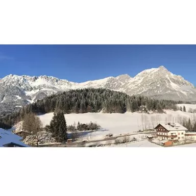 An impressive mountain landscape in winter with snow-covered peaks. In the foreground, wide fields and some buildings can be seen.