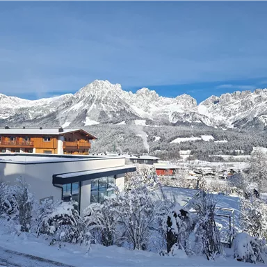 A snowy landscape with majestic mountains in the background. In the foreground, modern buildings and snow-covered trees stand.