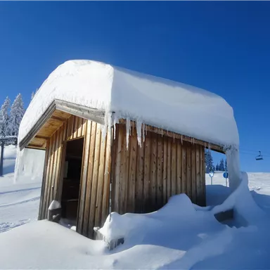 Eine kleine Holzhütte, bedeckt mit Schnee, steht unter einem klaren blauen Himmel. Rundherum ist die Landschaft mit frischem, unberührtem Schnee bedeckt.