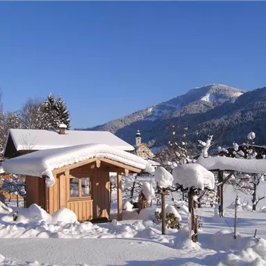 Ein gemütliches Holzhaus im Schnee mit schneebedeckten Landschaften und Bergen im Hintergrund. Der strahlend blaue Himmel vervollständigt die winterliche Szene.