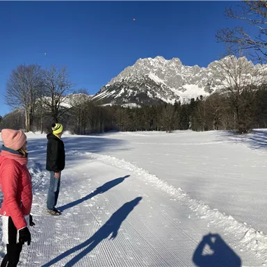 Zwei Personen stehen auf einem schneebedeckten Feld und blicken auf Berge im Hintergrund. Der Himmel ist klar und blau, was eine helle winterliche Atmosphäre schafft.