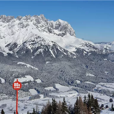 A wintry mountain landscape with snow-covered peaks and a clear blue sky. In the foreground, there is a signpost with a speed designation.