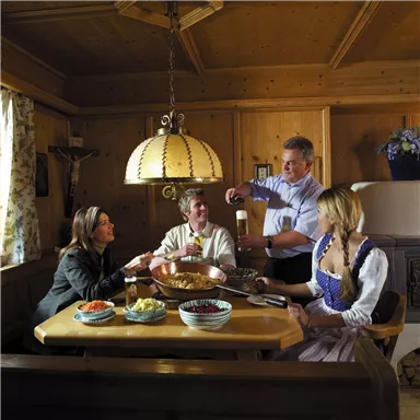 Eine gemütliche Runde am Tisch in einem traditionellen Holzraum. Die Gäste genießen Essen und Trinken in bayerischer Tracht.