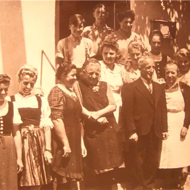 Eine Gruppe von Männern und Frauen steht zusammen vor einem Gebäude. Die Personen tragen verschiedene traditionelle und formelle Kleidungsstücke.