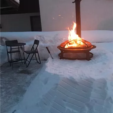 A cozy fire in a snow pile, surrounded by tall snow walls. In the background, there are two chairs and part of a house.