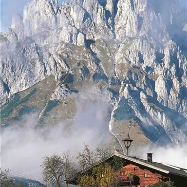 An impressive mountain landscape with high, rocky peaks and a picturesque alpine meadow in the foreground. The sky is partially cloudy and the surroundings appear peaceful.