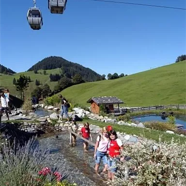 Eine malerische Landschaft mit grünen Wiesen und Bergen im Hintergrund. Menschen gehen durch ein Wasserbecken, während Seilbahnen darüber schweben.