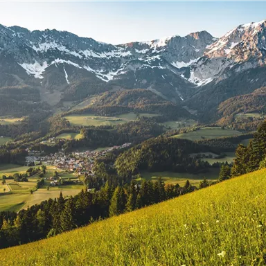 Eine beeindruckende Berglandschaft mit schneebedeckten Gipfeln und grünen Wiesen. Im Tal liegt ein kleines Dorf, umgeben von der Natur.