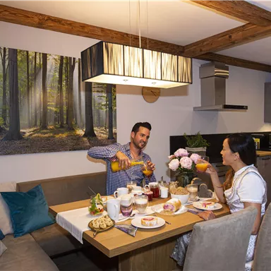 A cozy dining area with a wooden table, set with a breakfast of pastries, jam, and fruit. Two people are enjoying a shared breakfast and toasting with drinks.