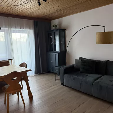 Ein gemütliches Wohnzimmer mit einem grauen Sofa und einem Holz-Esszimmertisch. Große Fenster sorgen für viel Tageslicht und eine entspannte Atmosphäre.