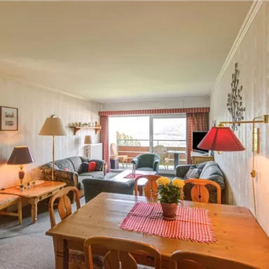 A comfortable living room with cozy furniture and plenty of natural light. The room features a dining table, plants, and a view of the balcony.
