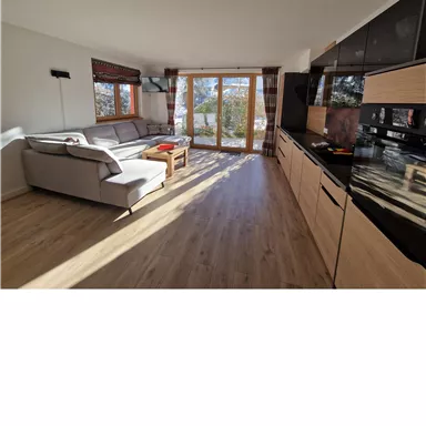 A modern living room with large windows and plenty of natural light. There is a comfortable sofa and a kitchenette.