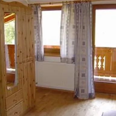 A cozy room with a wooden wardrobe and curtains. A lot of daylight can be seen through the windows.