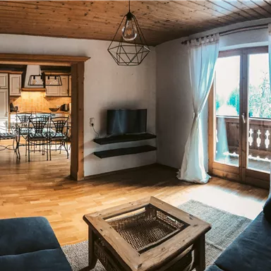 Ein gemütliches Wohnzimmer mit Holzdecke und großen Fenstern. Im Hintergrund ist ein Essbereich mit einem Tisch und Stühlen sichtbar.