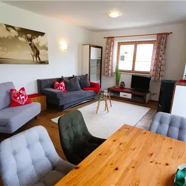 A bright living room with modern furniture and a large window. There is a cozy sofa, a wooden dining table, and a TV corner.