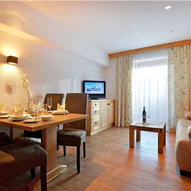 Ein gemütliches Wohnzimmer mit einem Essbereich und einem Fernseher. Die Fenster sind mit Vorhängen dekoriert und es gibt ein Sofa sowie einen Couchtisch.