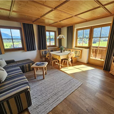 Ein gemütliches Wohnzimmer mit Holzmöbeln und Panoramafenstern. Es gibt ein Sofa, einen kleinen Tisch und einen Fernseher, sowie einen Blick auf die Berge.