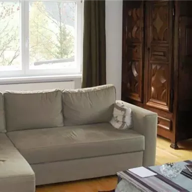 A cozy living room with a beige sofa and a large window. In the background, there is a beautiful dark cabinet.