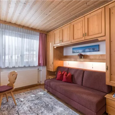 Ein gemütliches Wohnzimmer mit Holzvertäfelung und einem roten Sofa. Der Raum hat große Fenster, die viel Tageslicht hereinlassen.