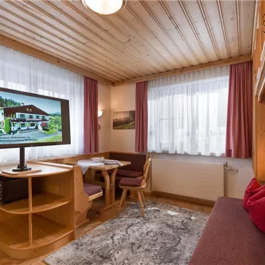 Ein gemütlicher Wohnraum mit Holzverkleidung und großen Fenstern. Ein Tisch und Stühle sowie ein Fernseher runden die Einrichtung ab.