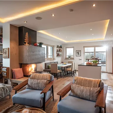 Ein modernes, stilvolles Wohnzimmer mit einem gemütlichen Kamin. Der Raum ist hell erleuchtet und hat eine offene Verbindung zur Küche und einer Esstisch-Ecke.