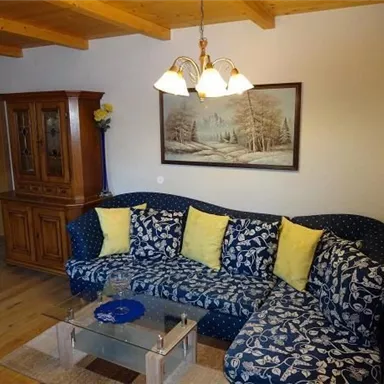 Ein gemütliches Wohnzimmer mit einem blauen Sofa, auf dem gelbe Kissen liegen. An der Wand hängt ein Bild einer Winterlandschaft und ein schöner Holztisch steht im Raum.
