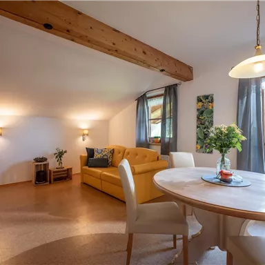 Ein gemütlicher Wohnraum mit einem gelben Sofa und einem Esstisch. Große Fenster lassen viel Licht herein und bieten einen Blick auf die Natur.