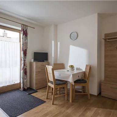 Ein gemütlicher Wohnraum mit einem Esstisch, Stühlen und Fernseher. Große Fenster sorgen für viel Tageslicht.